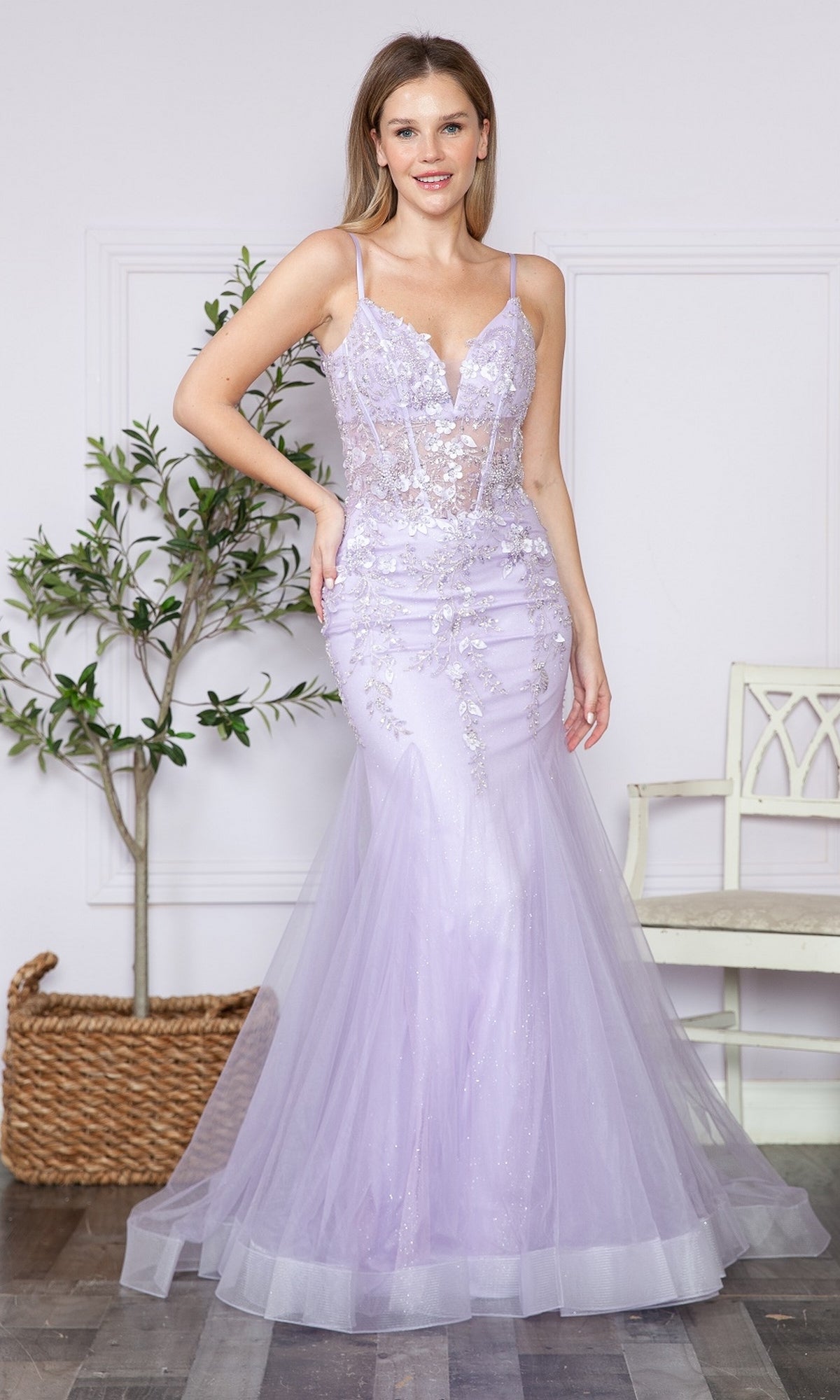 A woman stands indoors wearing the Sheer-Corset-Bodice Long Mermaid Prom Dress 9374 in lavender. Sheer panels and floral embroidery accent the fitted gown, with a potted plant and a white chair in the background.